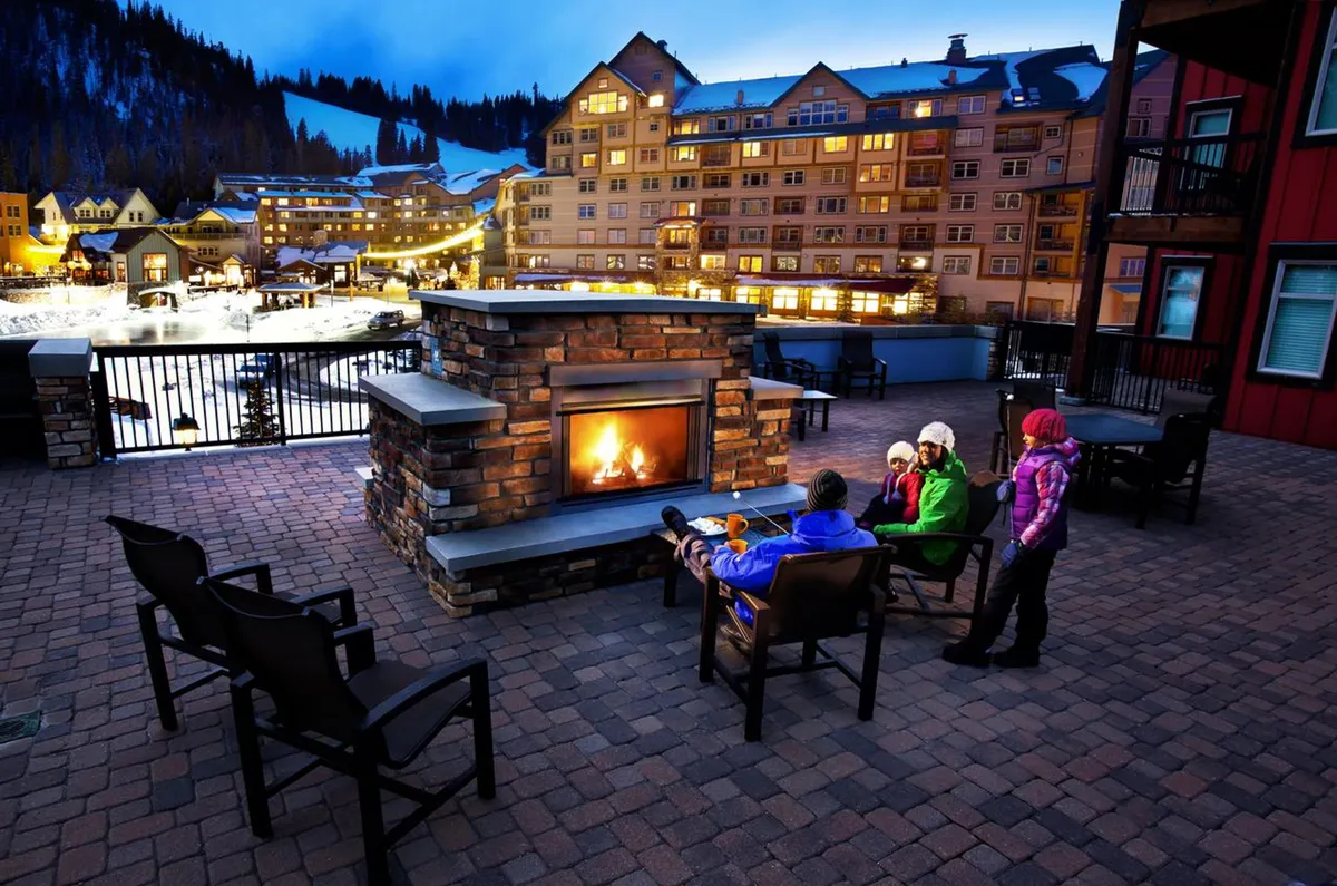 An inviting outdoor fireplace and seating area with a stunning mountain view, adjacent to the lodge’s facade. The scene is set for socializing, warming up, or unwinding after a day in the snow.