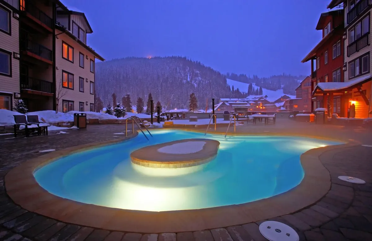 A stunning outdoor hot tub area surrounded by snow, with the mountains visible in the background. Steam rises off the water, suggesting warmth and relaxation even in winter. The scene hints at evening gatherings and peaceful stargazing, with the inviting glow of the hot tub contrasting beautifully with the snowy landscape.