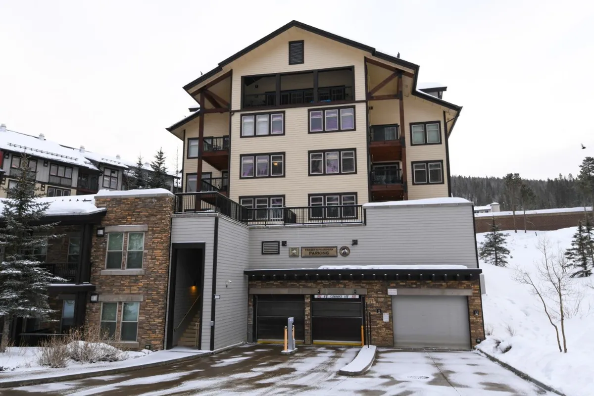 A welcoming multi-story resort condo building with a stone facade, garage entrance, and snow-covered ground, suggesting immediate access to winter activities. The exterior feels inviting and blends natural materials with modern comfort, reflecting a premium mountain resort setting.