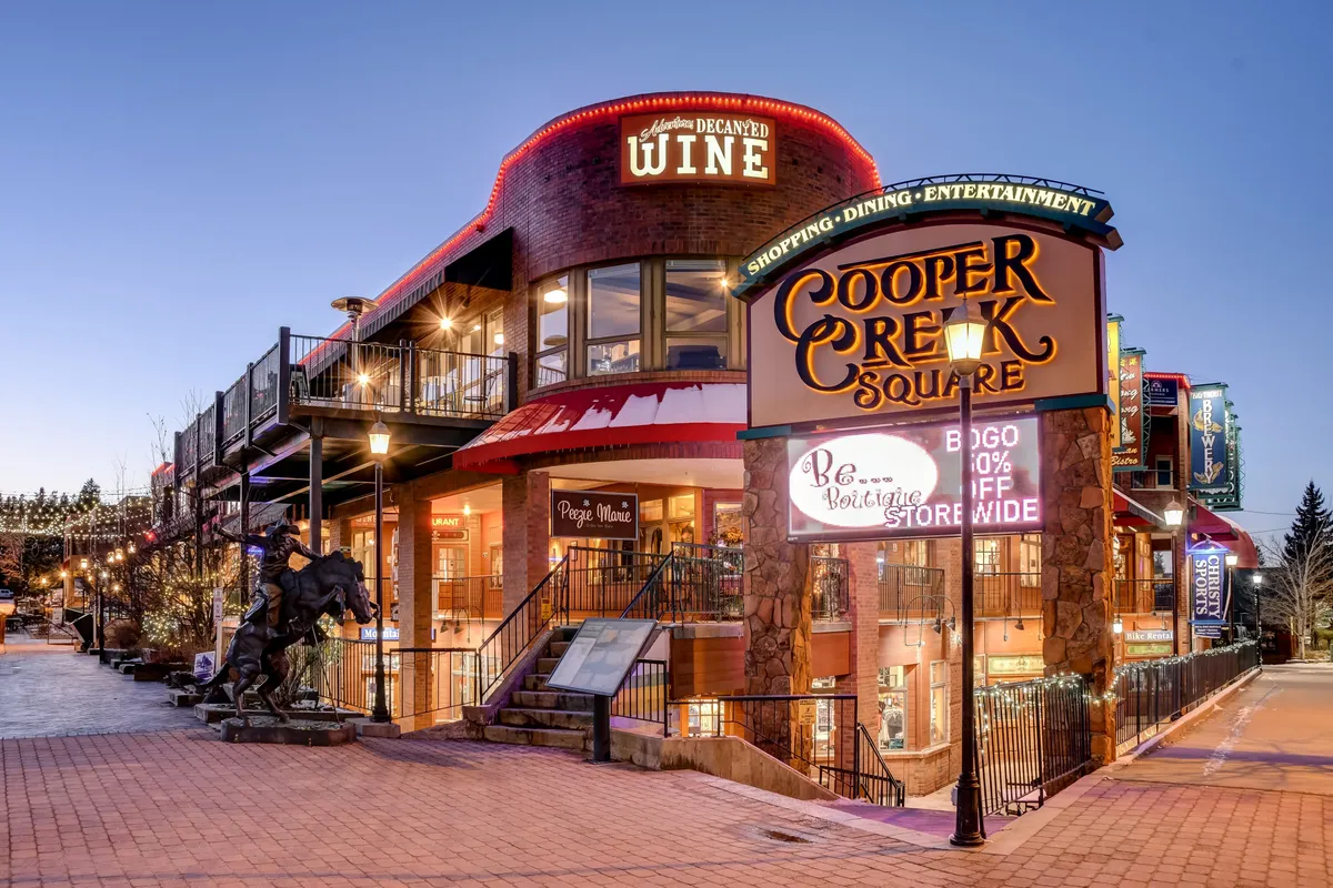A festive street scene at Cooper Creek Square, featuring decorative lighting, a wine shop entrance, a unique sculpture, and hints of bustling activity. The exterior is lively and inviting, ideal for shopping, dining, and entertainment.