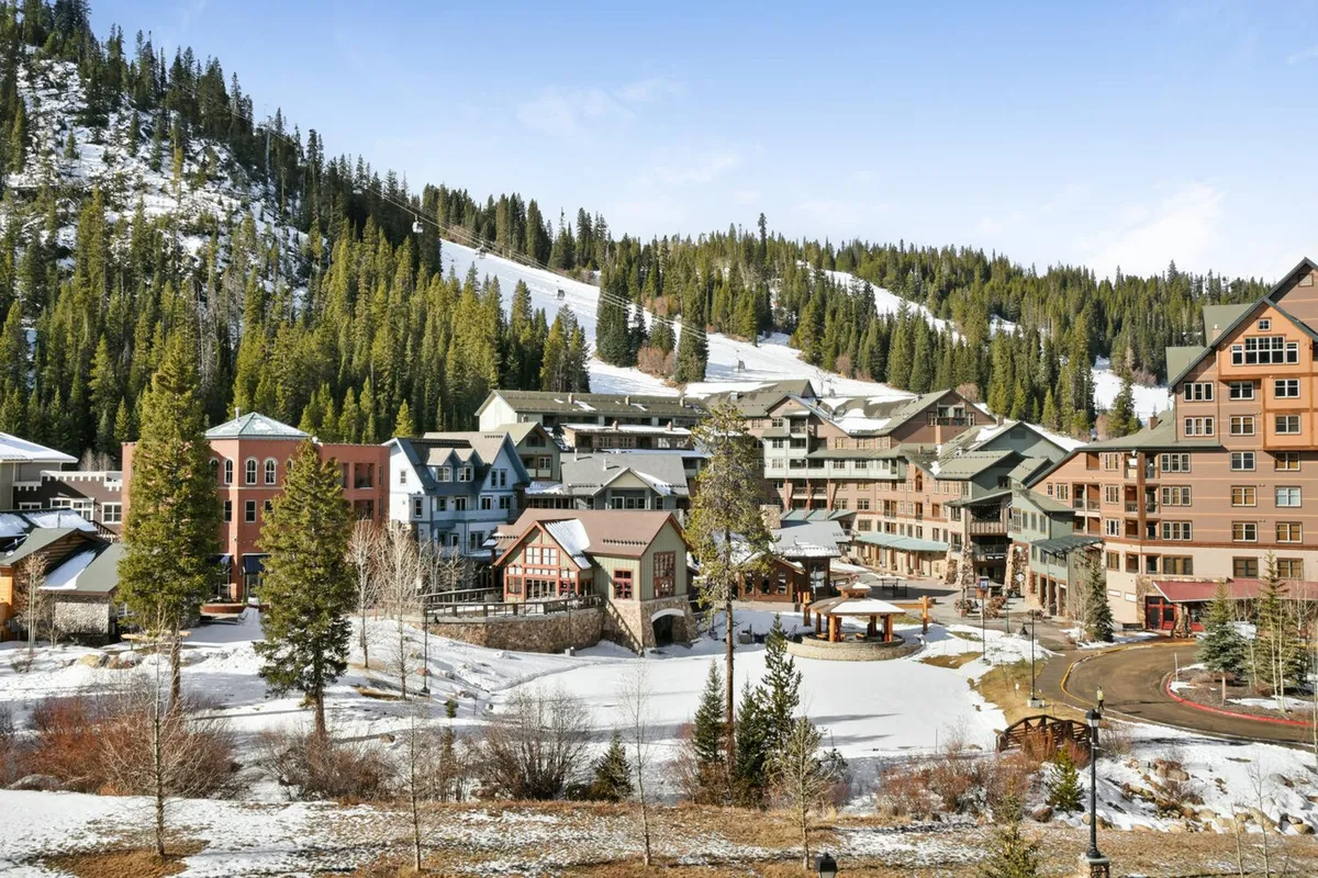A breathtaking view of snow-covered mountains, colorful resort village buildings, pine trees, and open space. The scene radiates adventure and serenity, capturing the surrounding environment and the vibrant resort atmosphere.
