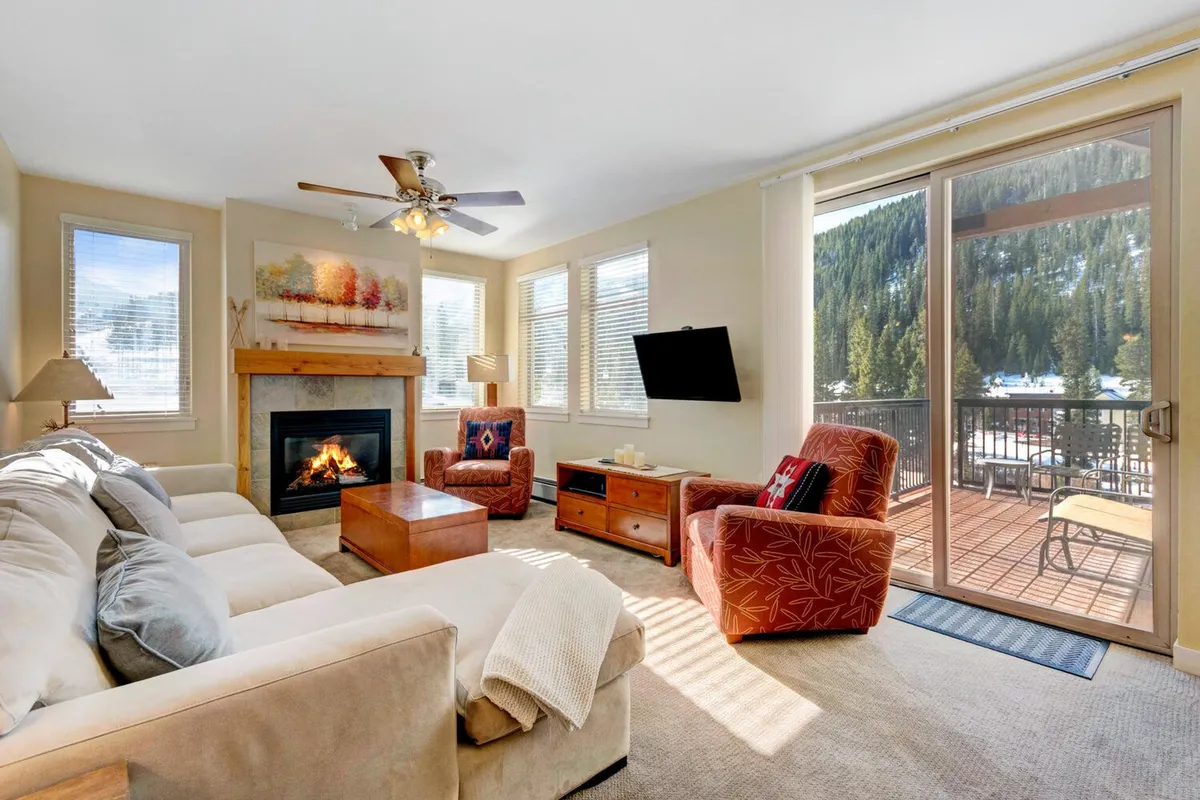 A warmly lit living room featuring a rustic gas fireplace as its centerpiece, flanked by plush seating and a modern coffee table. Large windows frame expansive mountain views, and a glass door leads directly to a private deck, reinforcing the connection to nature and the sense of seclusion. The overall arrangement is open-concept, with clear sightlines from the living to the deck area.