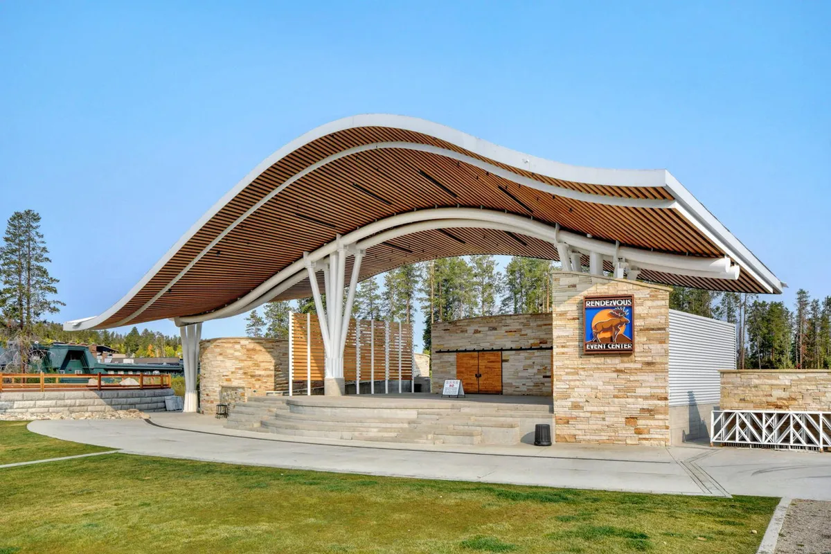 This exterior image showcases a curved roof structure with a stone façade and wooden accents, likely representing a community or event venue. The scene suggests lively outdoor events, concerts, and community gatherings, capturing the festive and communal spirit of the area.