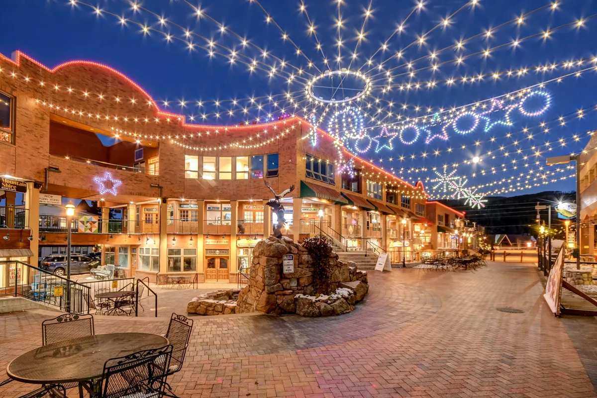 This exterior scene features a lively downtown area with string lights overhead, a decorative stone fountain, a brick walkway, and shops with illuminated windows. People can be seen strolling, dining outdoors, and shopping, capturing the vibrant spirit of the nearby town.