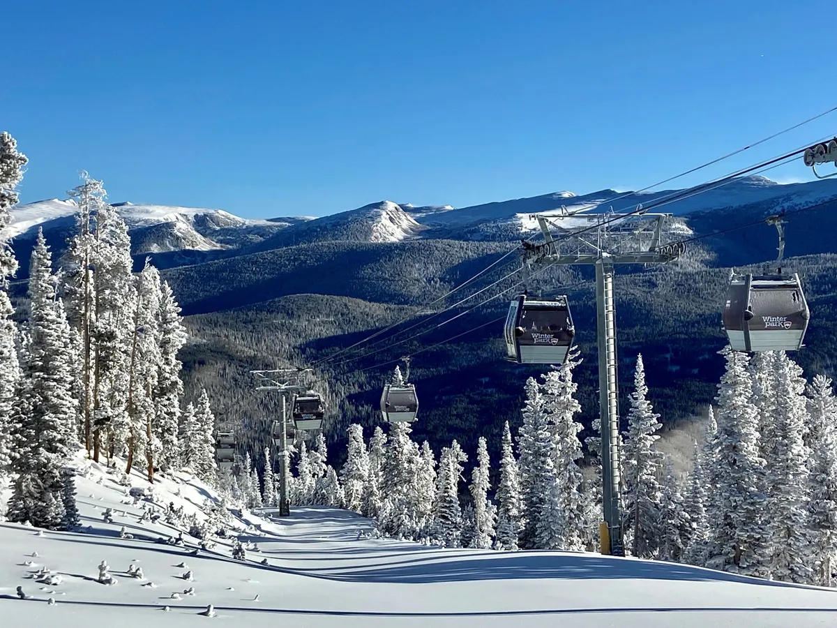 A panoramic winter view of a ski lift gliding above snow-covered trees, set against a dramatic mountain range. The fresh powder and lift hint at immediate access to ski runs and snowboarding terrain.