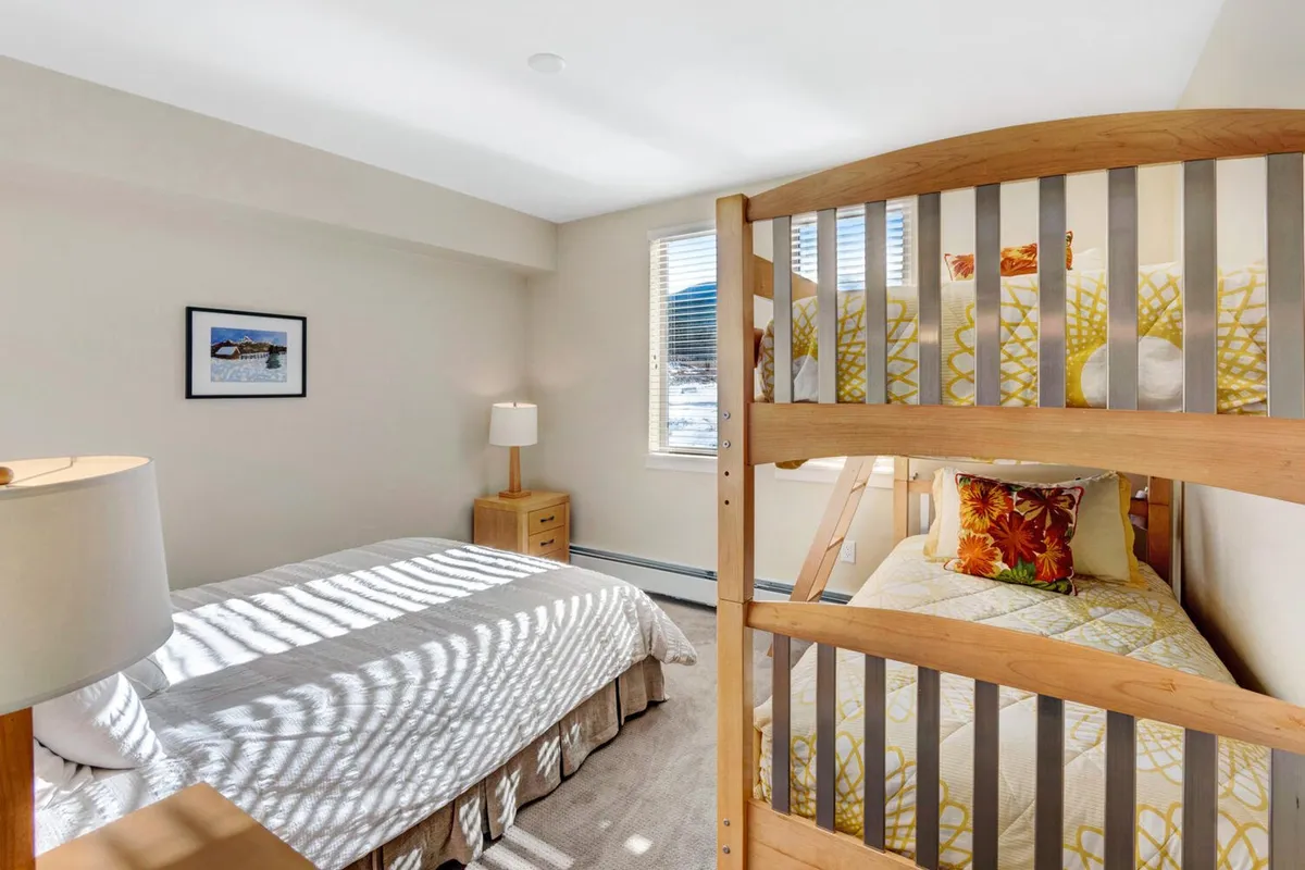 This image depicts a welcoming second bedroom featuring a queen bed and a twin bunk bed. Soft lighting, playful yet sophisticated décor, and a window with natural light create a comfortable setting. The arrangement is perfect for families, encouraging both restful sleep and playful bonding moments.