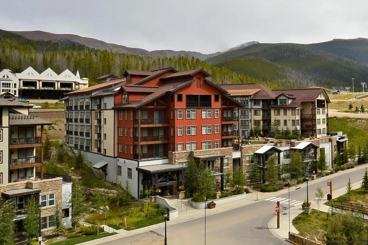 The image showcases a multi-story, lodge-style building surrounded by lush trees and a dramatic mountain backdrop. The atmosphere is cozy yet adventurous, with hints of exploration such as nearby trails and inviting outdoor spaces.
