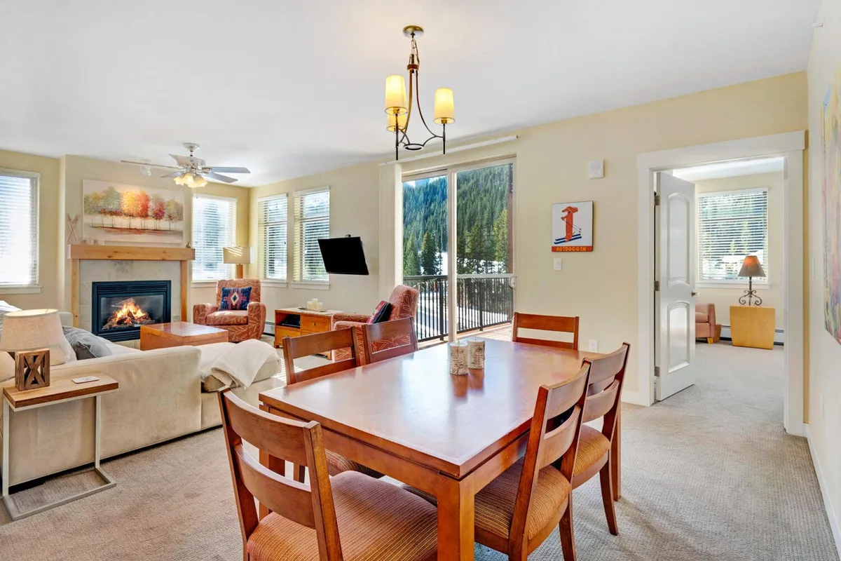 An inviting living and dining area with a prominent fireplace, elegant dining table, and expansive windows that showcase the mountain landscape. Artistic touches and comfortable seating make the space feel both upscale and homey.